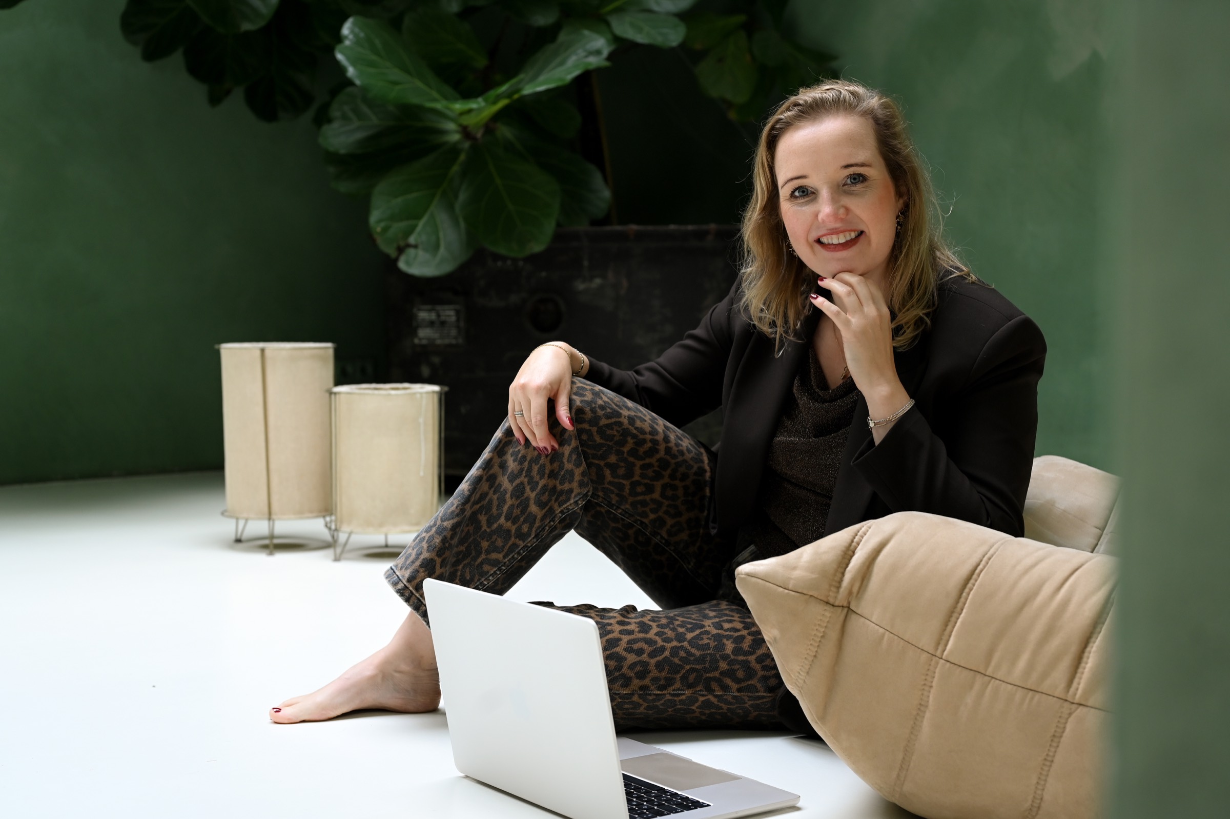 Selma zit op de grond tegen een kussen met haar laptop voor zich, in een ruimte met een groene wand.